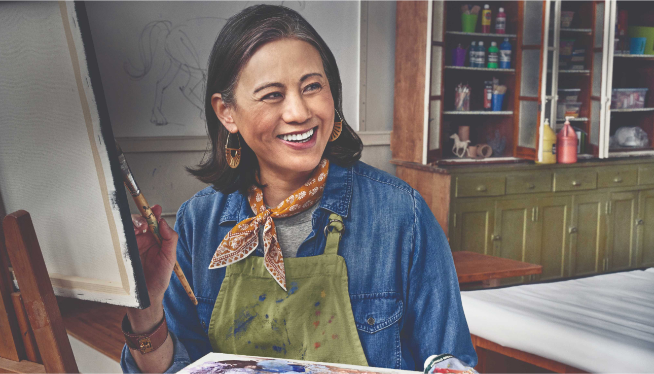 Older woman smiling while holding a paint brush in front of a canvas.