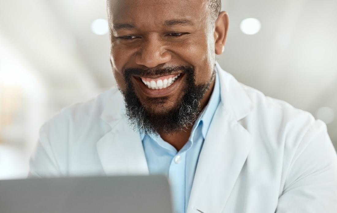 Smiling male eye care professional wearing a white coat and light blue shirt, looking at a laptop screen in a bright, modern setting