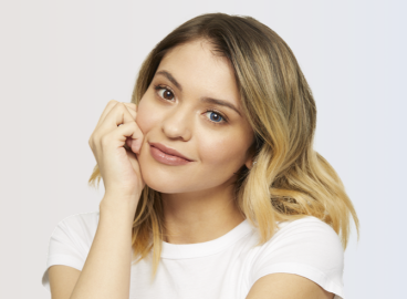 Smiling woman with blonde hair resting her chin on her hand, wearing a white top and one blue and one brown colored contact lens to show a side-by-side comparison effect.