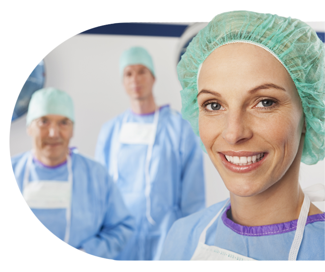 An image of a smiling surgeon wearing surgical scrubs with two other surgeons behind her smiling in the background of an operating room. 