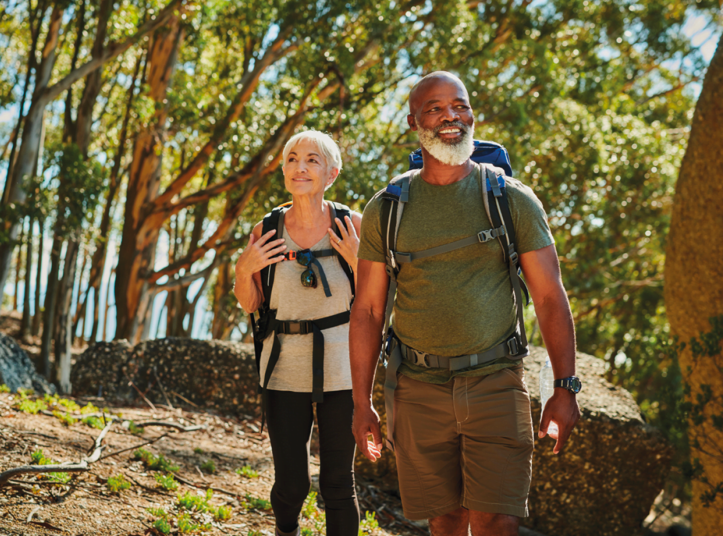 Un couple âgé en randonnée dans une forêt par une journée ensoleillée. La femme, aux cheveux blancs courts, porte un chandail clair, un pantalon noir et un sac à dos, tandis que l’homme, avec une barbe grise, porte un t-shirt vert, un short brun et un sac à dos. Ils ont l'air heureux et pleins d'énergie, profitant de l'aventure en plein air.