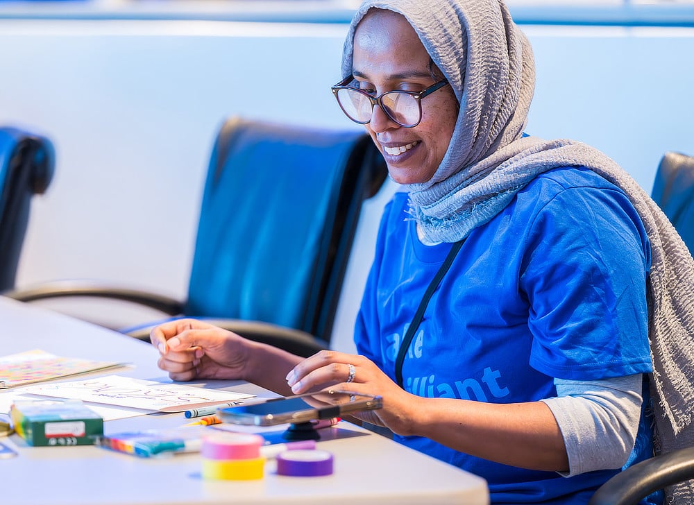 An Alcon volunteer wearing a headscarf and glasses is coloring a picture at a table. She is smiling and appears to be enjoying the activity.