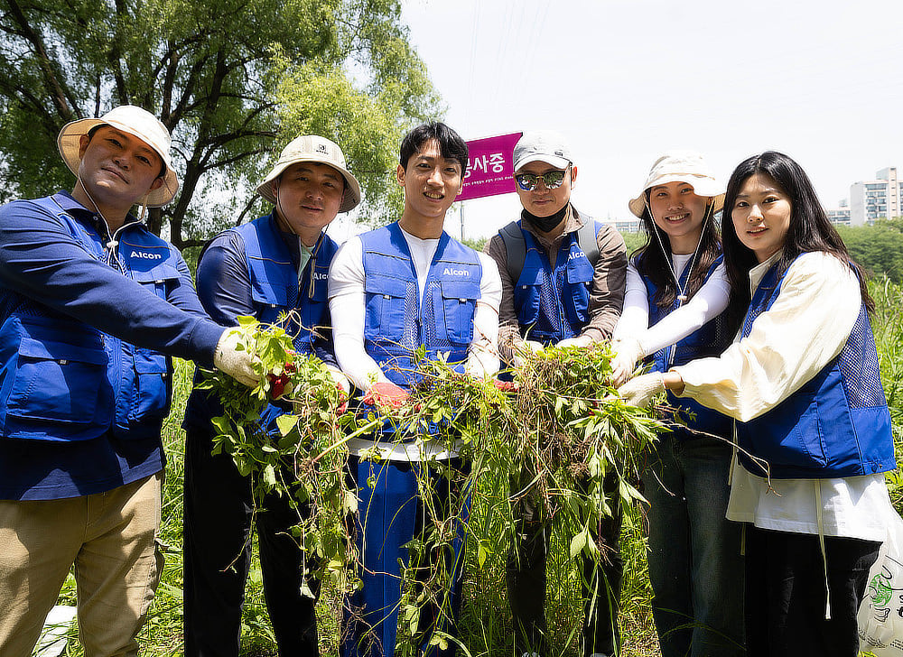 A group of Alcon volunteers wearing blue vests standing in a semi-circle and showing the weeds they have pulled.