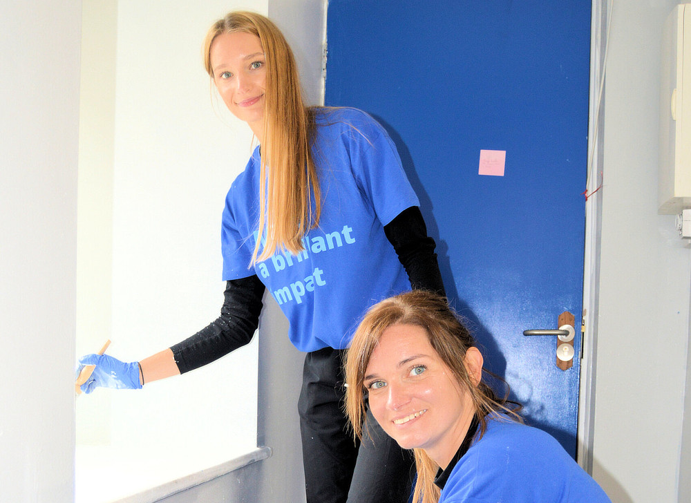 Two female Alcon volunteers are painting a wall white. They are wearing blue shirts and gloves, and one is kneeling while the other stands.