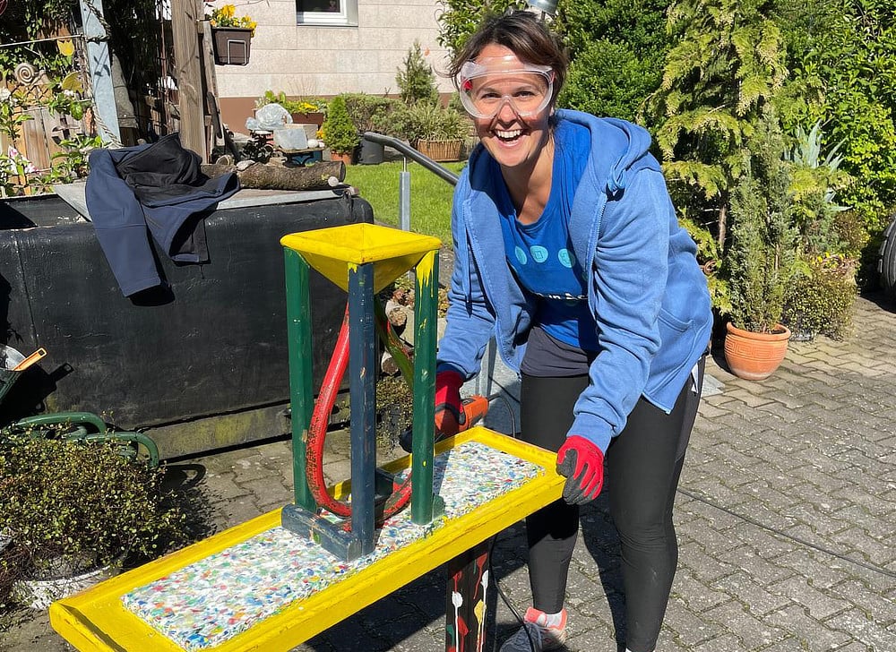 An Alcon volunteer is working on a brightly colored table in a garden. She is wearing gloves and safety glasses, and appears to be applying something to the tabletop.