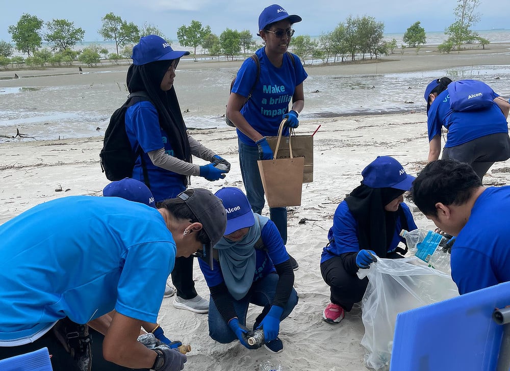 A group of Alcon volunteers wearing blue shirts picking up trash on a muddy beach. They are collecting plastic bottles and other debris.