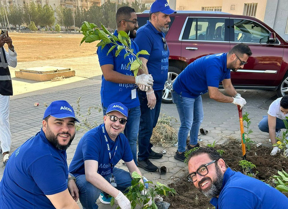 A group of Alcon volunteers wearing blue shirts are planting small trees in a garden bed. They are smiling and working together.
