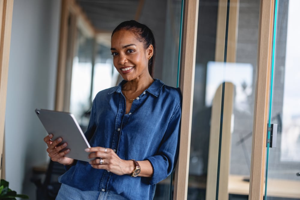 A smiling woman is looking down at a tablet she is holding in both hands. She is wearing a blue button-down shirt and a watch.