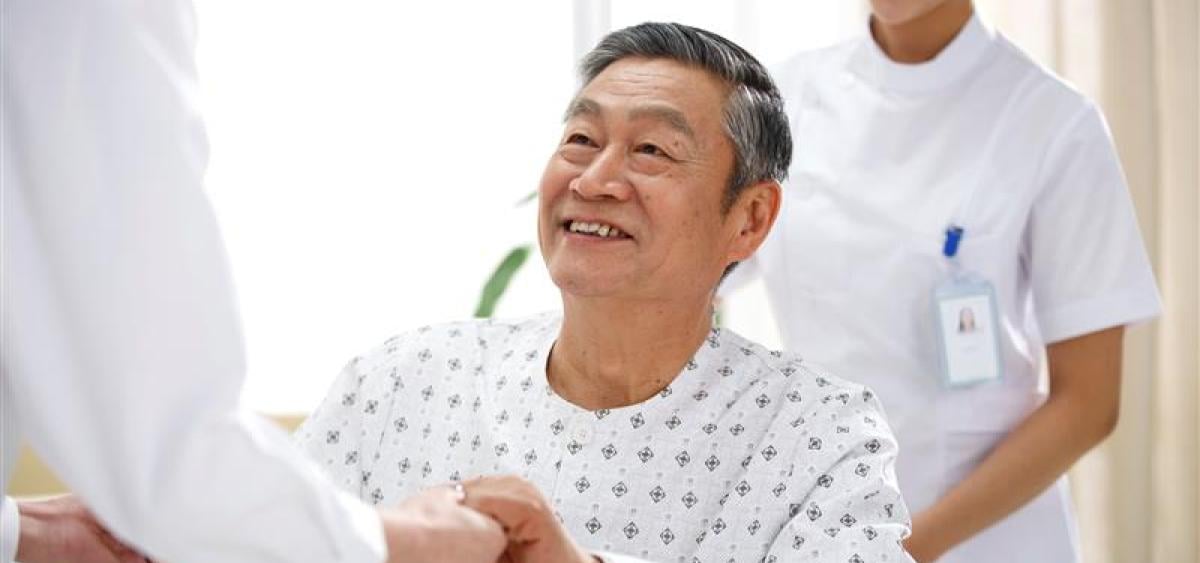 Smiling man in hospital gown grasping hands with doctor with nurse smiling behind him